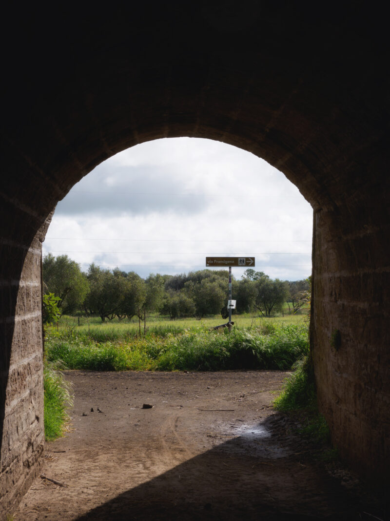 Cartello segnaletico della via Francigena visto da sotto un arco in pietra