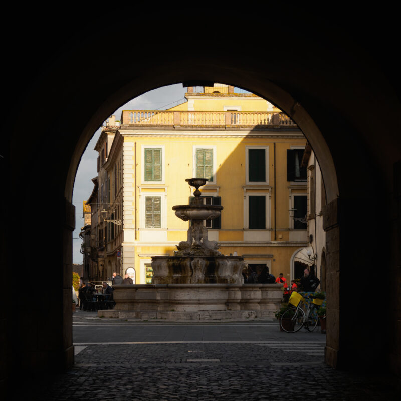Fontana barocca in una piazzetta vista dall'interno di un arco in pietra