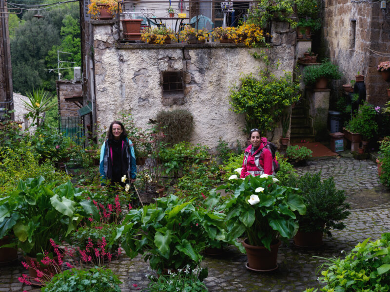 Due pellegrine con zaino in un piccolo giardino fiorito ricavato tra edifici storici