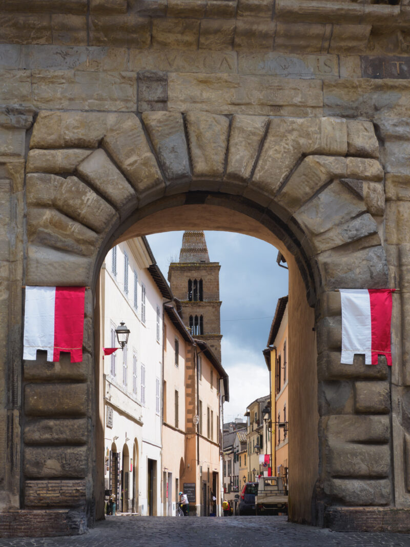 Porta medievale in pietra con bandiere rosse e bianche che introduce al centro storico di capranica