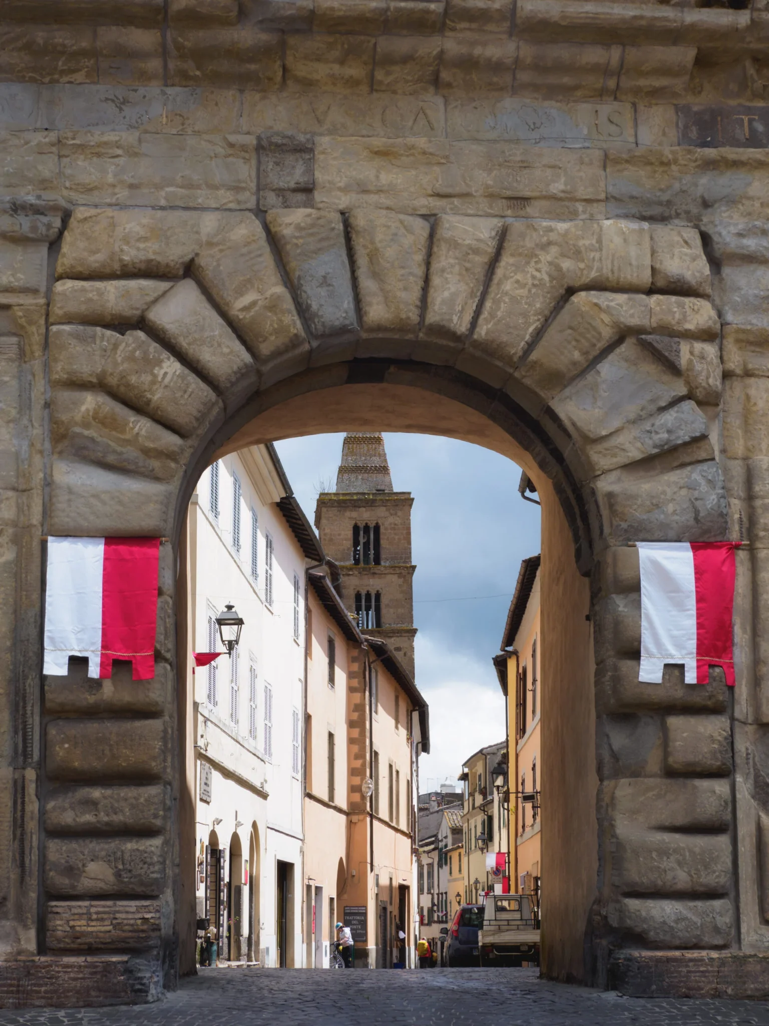 Porta medievale in pietra con bandiere rosse e bianche che introduce al centro storico di Capranica